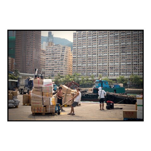 Workers move pallets of cargo on a pier in Hong Kong with tall buildings in the background. 