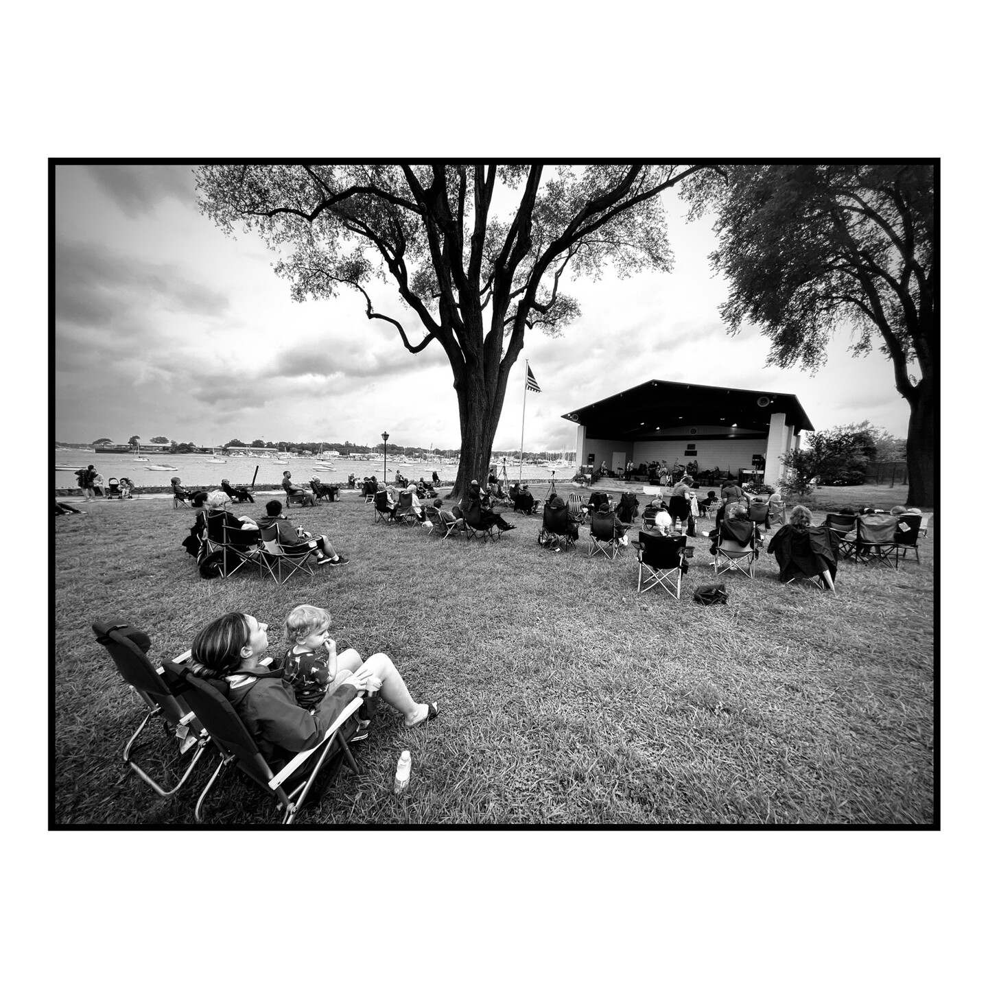 Black and white photo of people attending a concert on a lawn by the water.