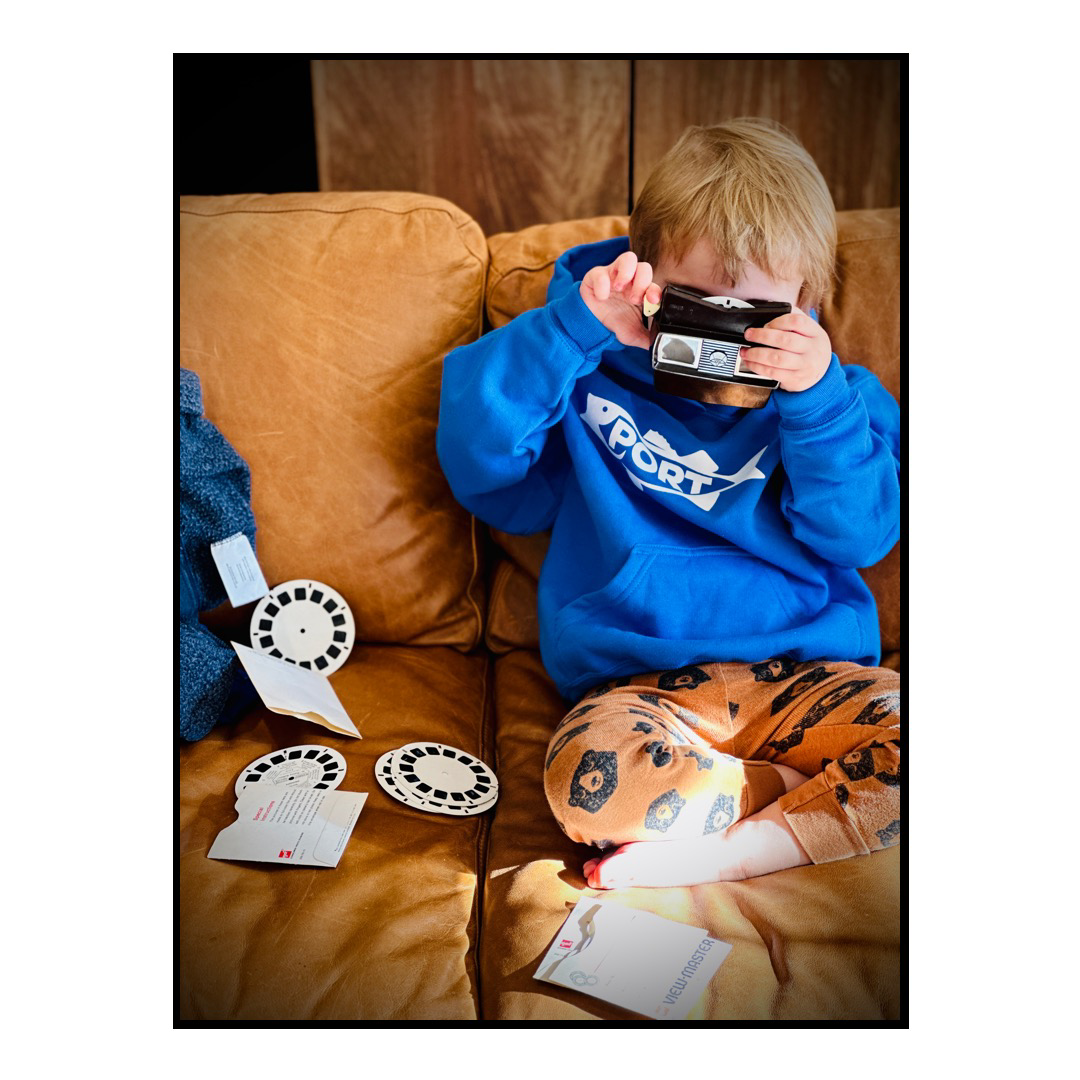 Son wearing a blue sweatshirt and sitting on a brown couch and playing with an old view master toy.
